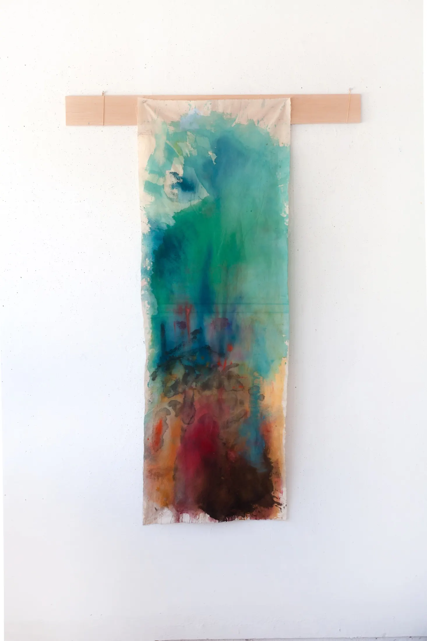 A long vertical dyed fabric banner hangs from a wooden batten on a white wall, shifting from teal and green at the top to red, ochre and dark brown at the bottom.