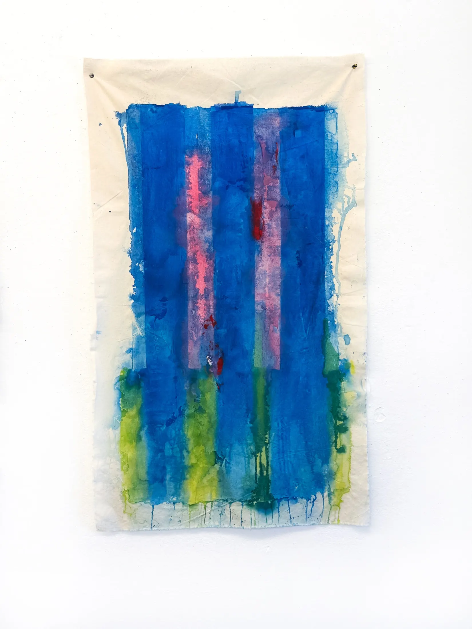 Vertical unstretched canvas pinned to a white wall, with a blue field, pink vertical bands, and green-yellow washes bleeding downward.