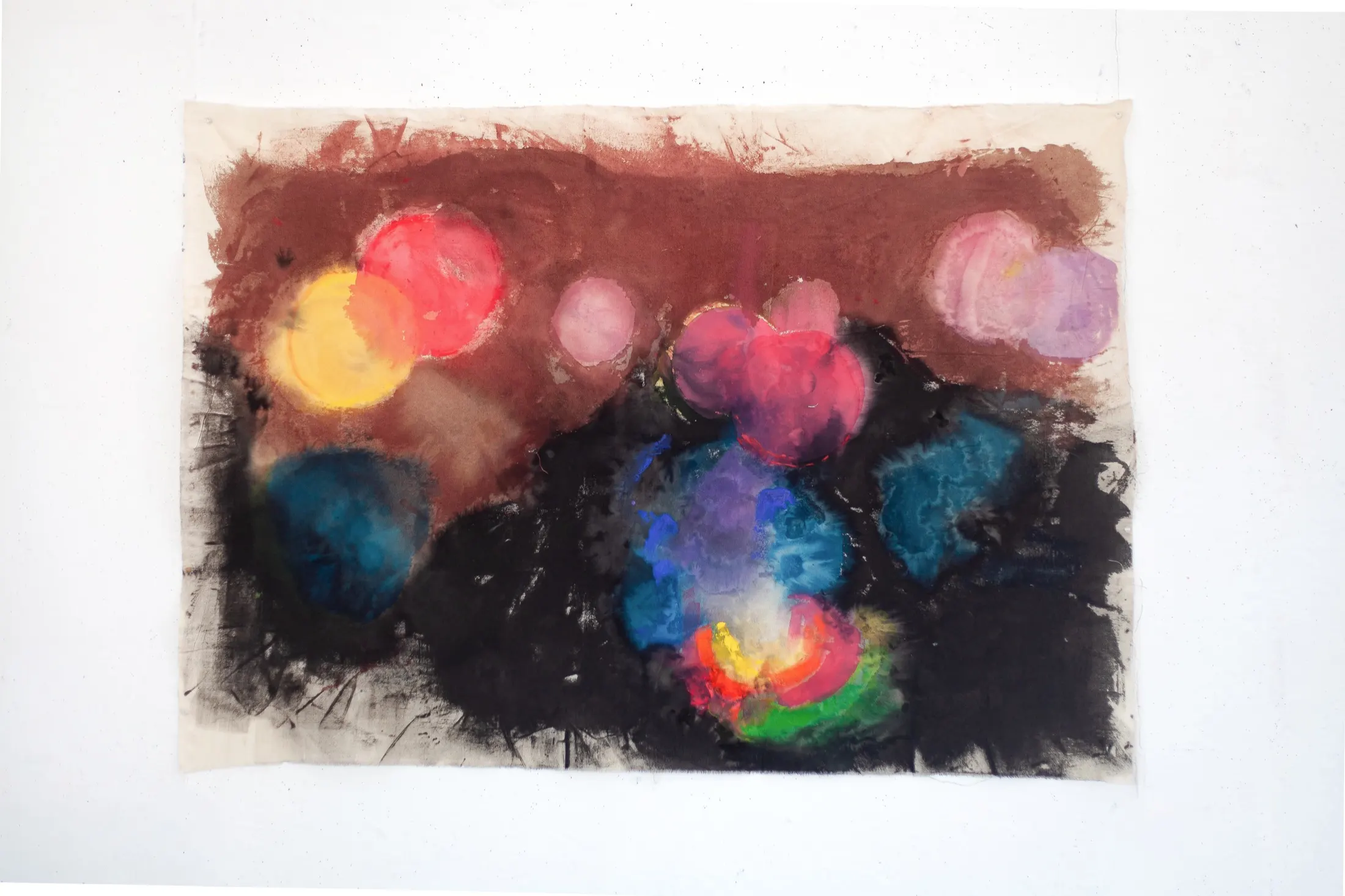 A horizontal raw-canvas fabric pinned flat to a white wall, with glowing circular blobs of red, yellow, pink and blue against maroon-brown and black grounds.