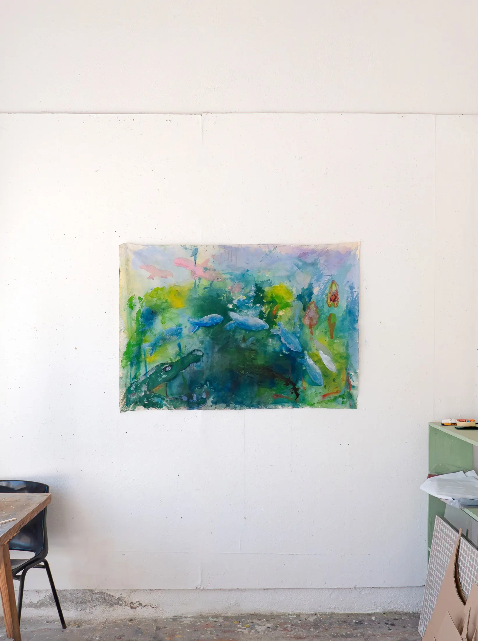 A landscape-format unstretched fabric painting of a pond-like scene with blue fish, green foliage and pink shapes hangs on a white studio wall.
