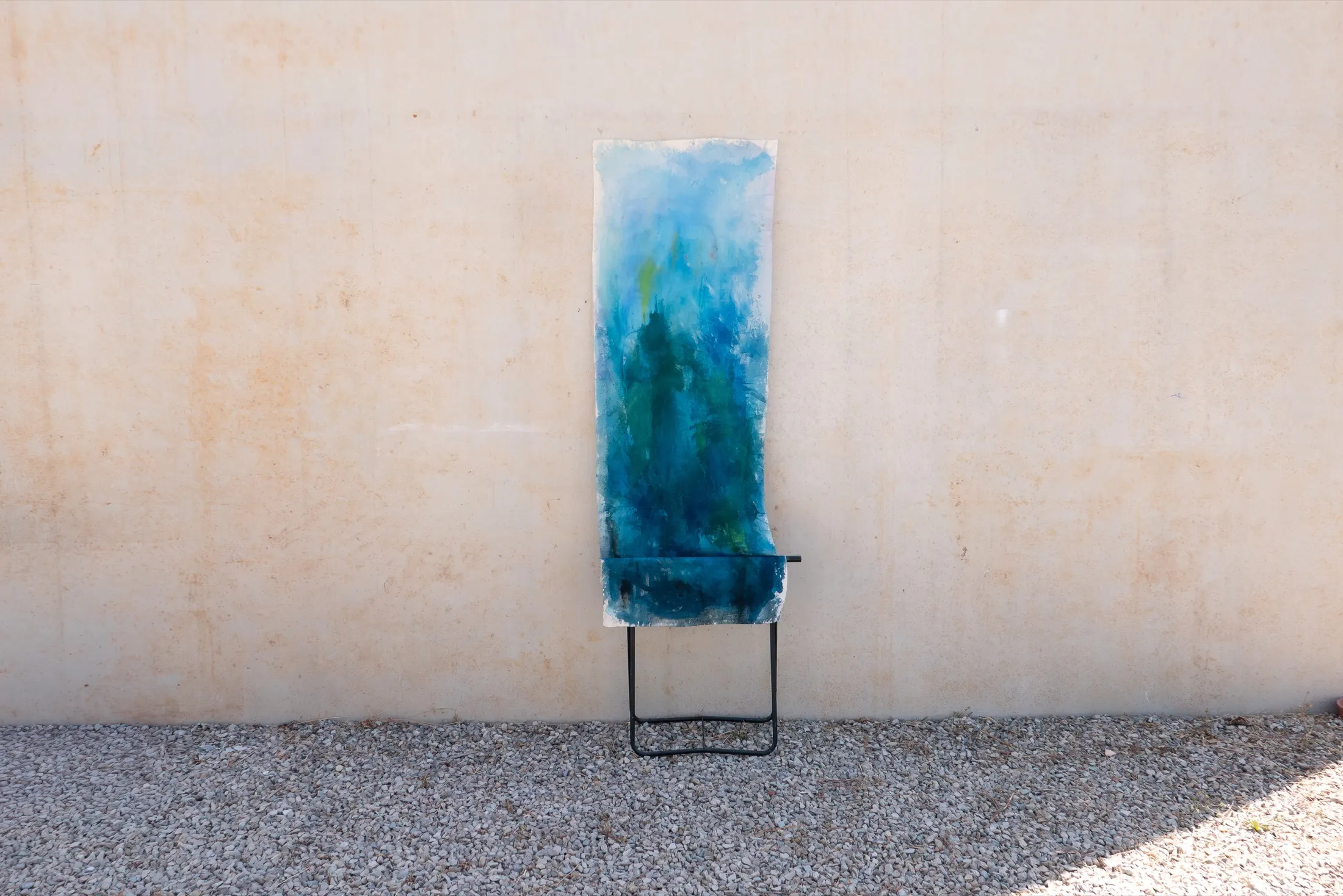 A tall blue-dyed fabric draped over a black metal folding stand leans against a beige outdoor wall above gravel.
